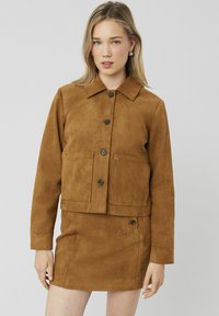 Veste en daim marron avec un col, fermeture à boutons à l'avant et deux grandes poches, associée à une mini-jupe en daim assortie.