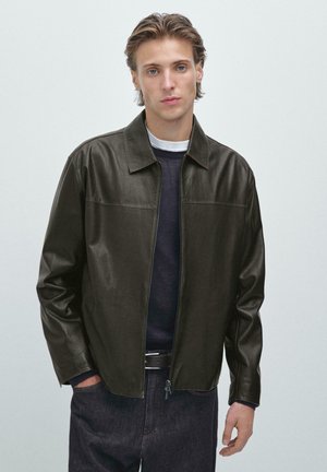 Dunkelgrüne Lederjacke, mit Reißverschluss vorne, Umlegekragen, lange Ärmel, glatte Textur, kombiniert mit einem marineblauen Pullover und Jeans.