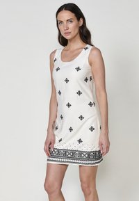 Ärmelloses weißes Kleid mit schwarzer Stickerei in Diamant- und Punktmustern, das einen detaillierten schwarzen Rand am Saum aufweist. Glatte Baumwolltextur.