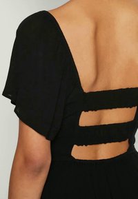 Melnā kleita ar īsām pūkainām piedurknēm un dziļu dekoltē aizmugurē, kurai ir izgriezumi gar muguru un struktūrēta auduma detaļas.