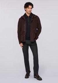 Giacca in suede marrone con chiusura a zip e bottoni, due tasche frontali, indossata su una maglia scura e abbinata a jeans scuri e scarpe marroni.