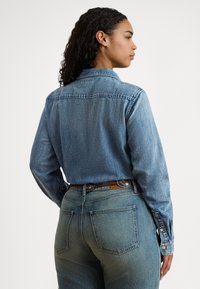 Žena s kučeravými vlasmi, oblečená v vybledlej modrej rifľovej košeli zastrčenej do rovnakých riflí s dekoratívnym hnedým opaskom, tvárou bočne.