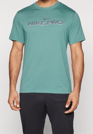 Hombre con camiseta de manga corta Nike Pro color verde azulado y pantalones negros, de pie contra un fondo claro y liso.