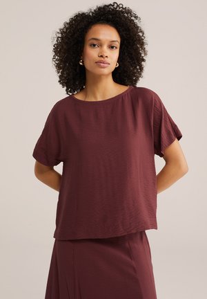 Femme aux cheveux bouclés portant des boucles d'oreilles créoles en or, un haut ample à manches courtes bordeaux et une jupe assortie, sur un fond uni.