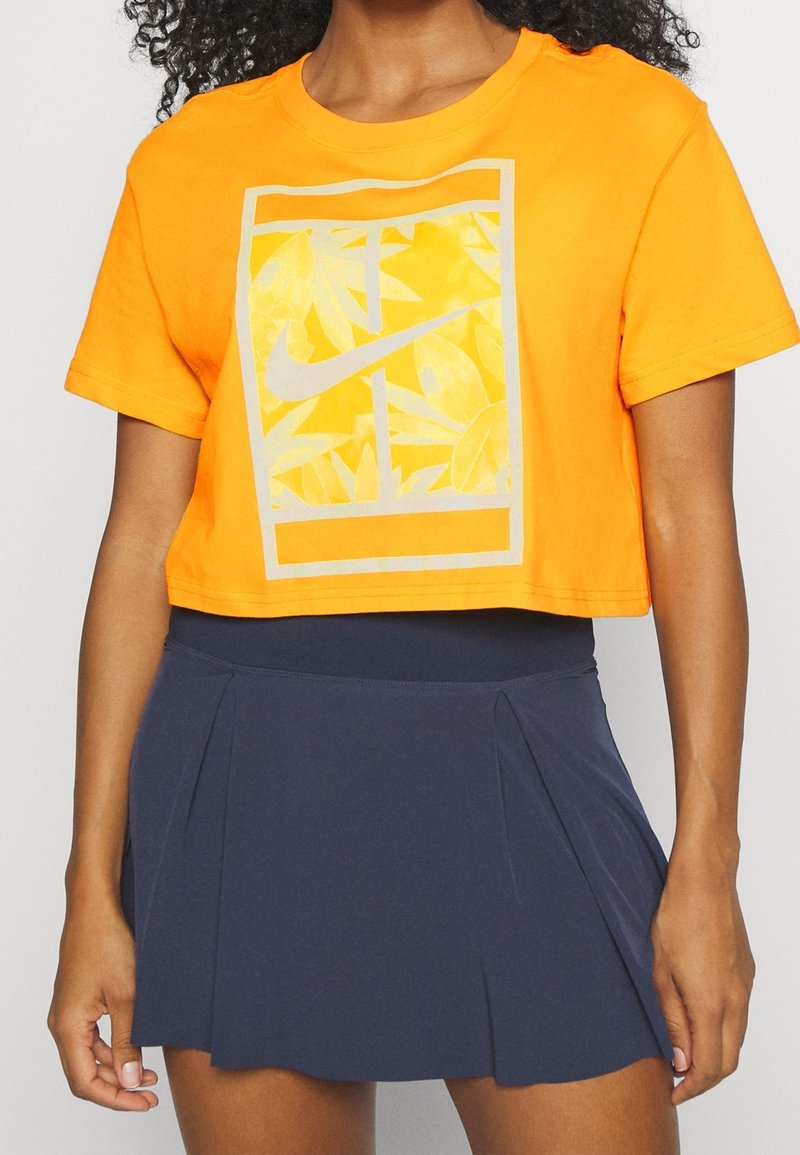 Bright orange cropped t-shirt with a textured yellow leaf pattern and gray Nike logo, paired with a navy blue pleated skirt.