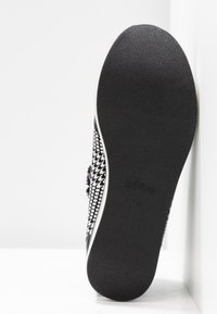 Semelle noire d'une chaussure à motif pied-de-poule noir et blanc, présentée debout contre un mur blanc d'angle.