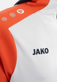 JAKO Sporttrikot mit einem weißen Oberteil, orangefarbigen und marineblauen Akzenten, strukturiertem Stoff und drei dekorativen Punkten auf der Schulter.