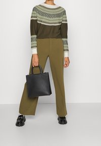 Olivgrüner Pullover mit geometrischem Muster, kombiniert mit olivgrünen, weit geschnittenen Hosen und einer schwarzen Lederhandtasche. Glänzende schwarze Schuhe runden das Outfit ab.