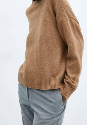 Personne portant un pull marron ample avec poignets et ourlet côtelés, associé à un pantalon gris clair, main dans la poche sur un fond uni.