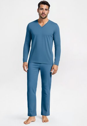 Uomo che indossa una maglietta a maniche lunghe blu con scollo a V e pantaloni abbinati, in piedi a piedi nudi contro uno sfondo bianco semplice.