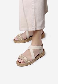 Beige espadrille sandaalit, joissa on kudottu päällinen, ristikkäiset elastiset hihnat ja juuttipohja. Litteä muotoilu, joka tuo esiin yksinkertaisen ja rentoja estetiikkaa.