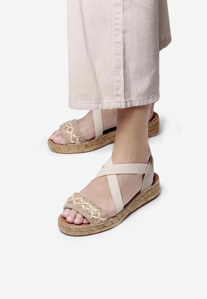 Beige espadrille sandaalit, joissa on kudottu päällinen, ristikkäiset elastiset hihnat ja juuttipohja. Litteä muotoilu, joka tuo esiin yksinkertaisen ja rentoja estetiikkaa.