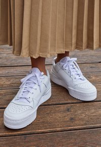 Vita lädersneakers med runda tår, perforerat mönster och platta vita snören. Sulan är texturerad och i plattformsstil, på trägolv.