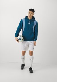 Felpa color teal con accenti azzurri e un logo, abbinata a pantaloni corti bianchi, scarpini neri e calze bianche. Tiene in mano un pallone da calcio.