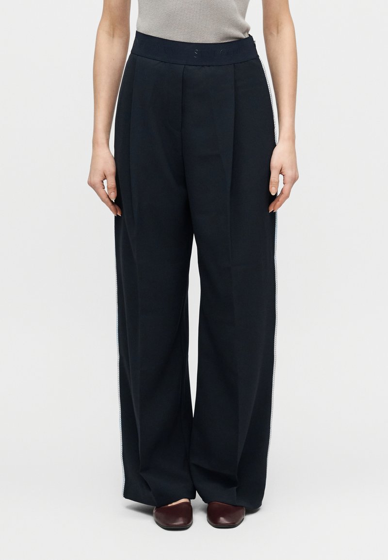 Wijde broekspijpen in donker marineblauwe stof met een hoge tailleband, subtiele plooien en zijaccenten met een contrasterende verticale bies.