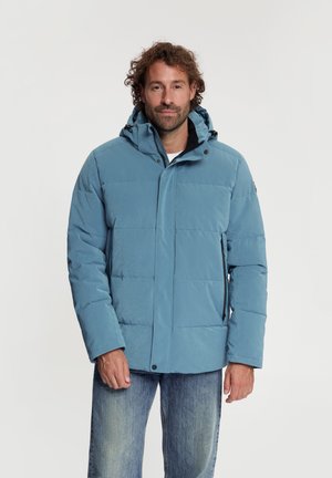 Man met krullend haar die een lichtblauw donsjasje met capuchon en een blauwe spijkerbroek draagt tegen een effen lichte achtergrond.