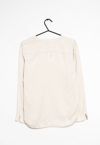 Camisa de manga larga en tejido beige claro con detalle en la parte posterior y aberturas en los laterales del dobladillo. Cuelga de un gancho negro sobre un fondo blanco.