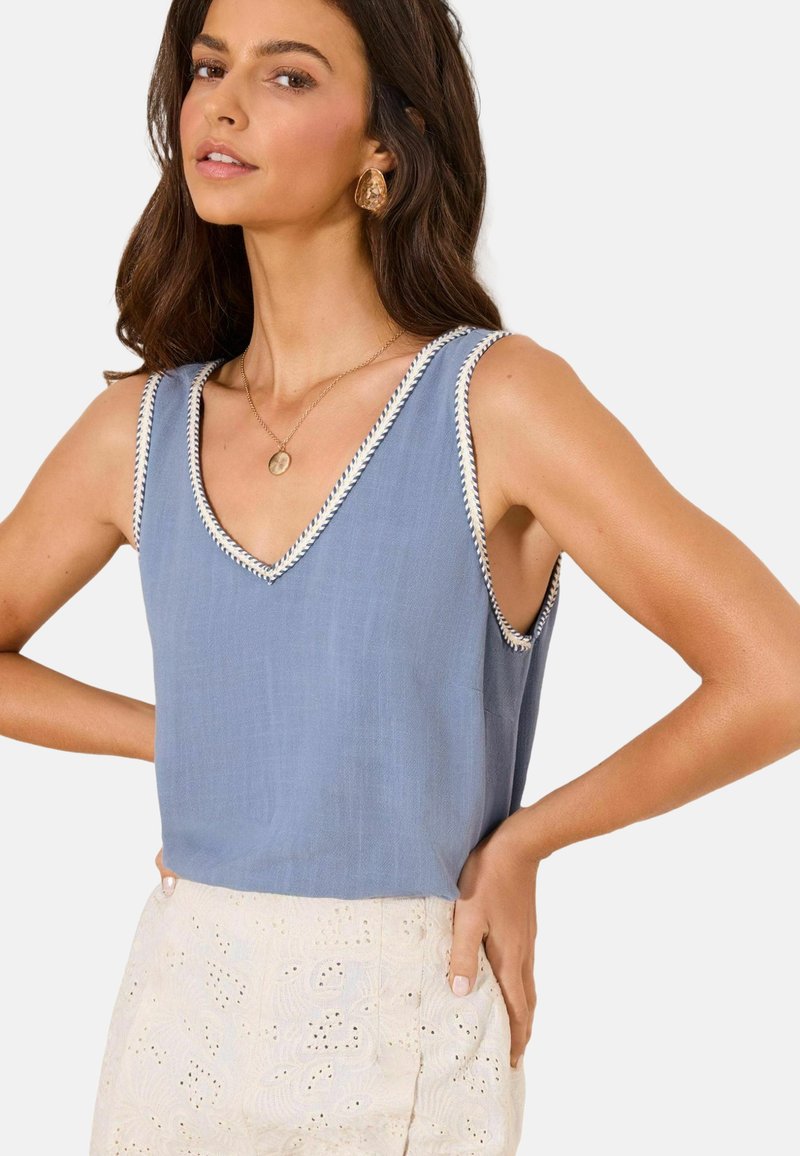 Mujer con top sin mangas azul claro con escote en V y ribete blanco, y falda de encaje color crema, con collar y pendientes con colgante dorado.