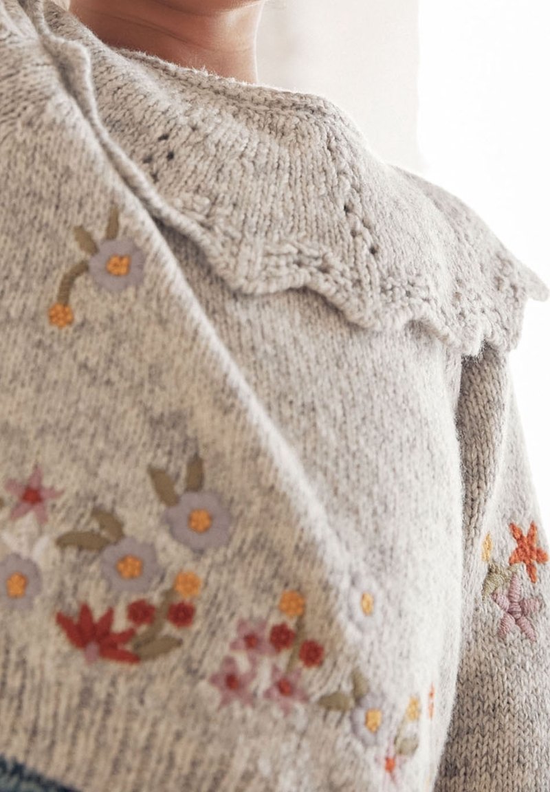 Gros plan sur un pull en maille beige avec un col en forme d'ondulations et une broderie florale aux couleurs douces et tamisées sur la poitrine et la manche.