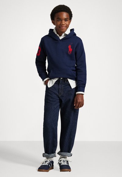 Jūras karakuģu stila kapuce ar sarkano polo logotipu un numuru 3, kombinēta ar sarullētām tumši zilām džinsām un jūras zila sporta apaviem ar baltām auklām.