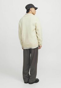 Hellbeige Oversized-Pullover mit einem ribbed Saum, über einem gestreiften Hemd getragen, kombiniert mit dunklen, weiten Hosen und schwarzen Schuhen.