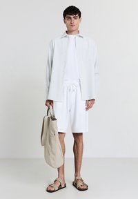 Uomo che indossa pantaloni corti bianchi, t-shirt bianca, camicia a righe chiare, sandali beige, mentre tiene una borsa a tracolla beige, in piedi contro uno sfondo bianco semplice.