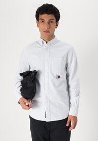Camisa de botones a rayas en algodón azul claro y blanco, con un bolsillo en el pecho y un parche de logo. Modelo sosteniendo un bolso negro.