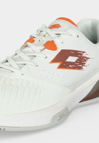 Hvid sportssko med orange accenter, hvide snørebånd, perforerede sider, et mønstret logo og tekstureret sål.