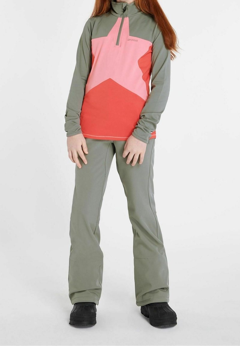 Personne portant un pantalon vert, des bottes noires et un haut à manches longues zippé avec des blocs de couleur géométriques roses, rouges et verts.