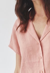Camicia in lino rosa chiaro con scollo a V, maniche corte e cinque bottoni frontali, che mostra una consistenza morbida e una vestibilità rilassata.