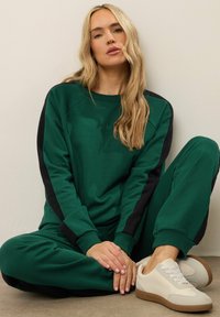 Pantaloni della tuta verdi e felpa abbinata con strisce laterali nere; materiale morbido; abbinati a sneakers beige con suola testurizzata.