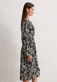 Schwarzblumenkleid mit langen Ärmeln und taillierter Taille. Besitzt weiße und gelbe Blumenmuster auf einem strukturierten Stoff. Midi-Länge.