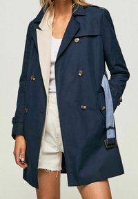 Mörkblå dubbelknäppt trenchcoat med en ljusfärgad krage, metallknappar och denimdetaljer på ärmarna.