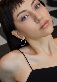Brinco em argola prateada com detalhes texturizados, usado por uma pessoa com cabelo liso e escuro, vestindo uma camisola preta contra um fundo quadriculado.