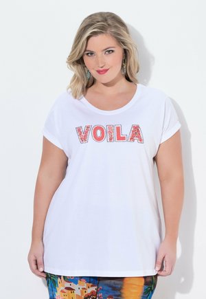 Kobieta z blond włosami do ramion, ubrana w białą koszulę z napisem „VOILA” oraz kolorowe spodnie we wzory, stojąca na białym tle.