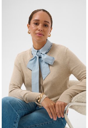 Femme au visage tacheté de rousseur portant un cardigan beige, un chemisier bleu avec un grand nœud, des boucles d'oreilles anneaux dorés et un bracelet en or, assise sur une chaise texturée.