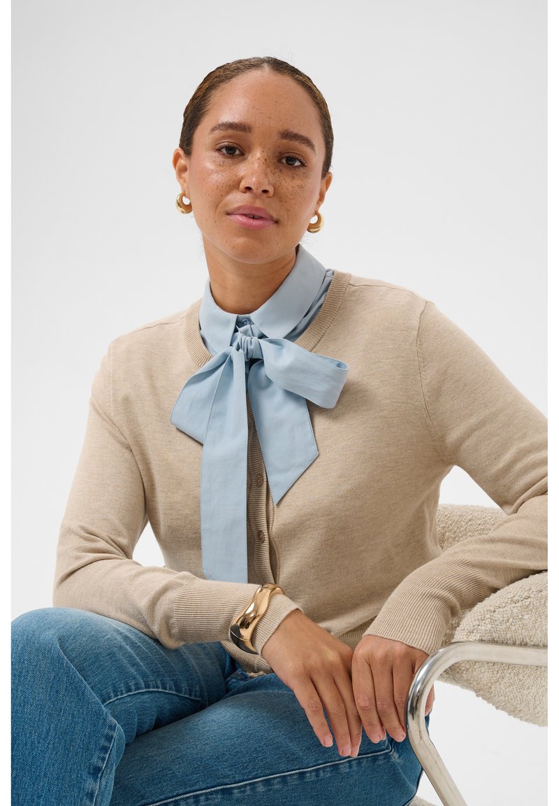 Femme au visage tacheté de rousseur portant un cardigan beige, un chemisier bleu avec un grand nœud, des boucles d'oreilles anneaux dorés et un bracelet en or, assise sur une chaise texturée.