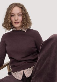 Brauner, gestrickter Pullover, der über ein gestreiftes Hemd mit Kragen getragen wird. Lockiges Haar, sitzt auf einem Holzstuhl, mit strukturierten Cord-Hosen.