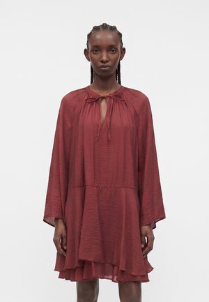 Modèle féminin aux cheveux tressés portant une robe ample, à manches longues, superposée, rouge rouille avec un col clé noué.