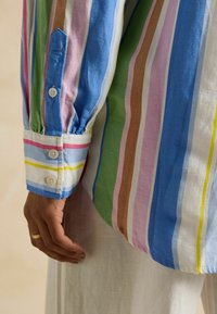 Mehrfarbig gestreiftes Hemd mit vertikalen Linien in Blau, Grün, Pink und Braun. Verfügt über knopfbare Manschetten und einen strukturierten Stoff. Hand in der Nähe des Saums gezeigt.
