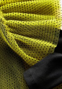 Tessuto giallo traslucido con pois neri, caratterizzato da pieghe e un polsino nero testurizzato. Il materiale appare leggero e arioso.