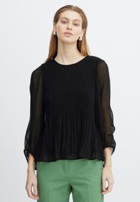 Zwarte geplooide blouse met doorzichtige mouwen en een ronde halslijn, met een vloeiende silhouet. Model gecombineerd met groene broeken.