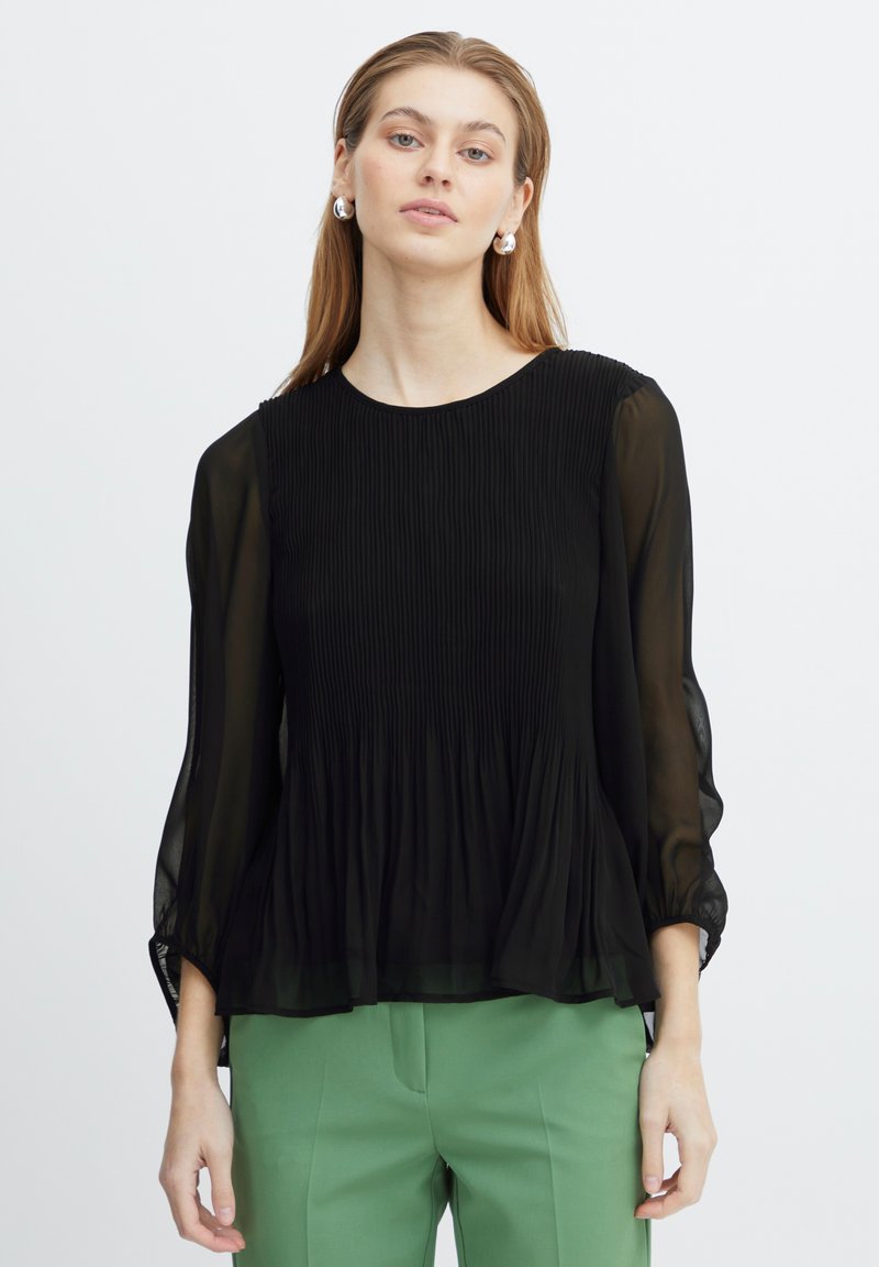 Bluză neagră plisată cu mâneci transparente și decolteu rotund, având o siluetă fluidă. Modelul a combinat-o cu pantaloni verzi.