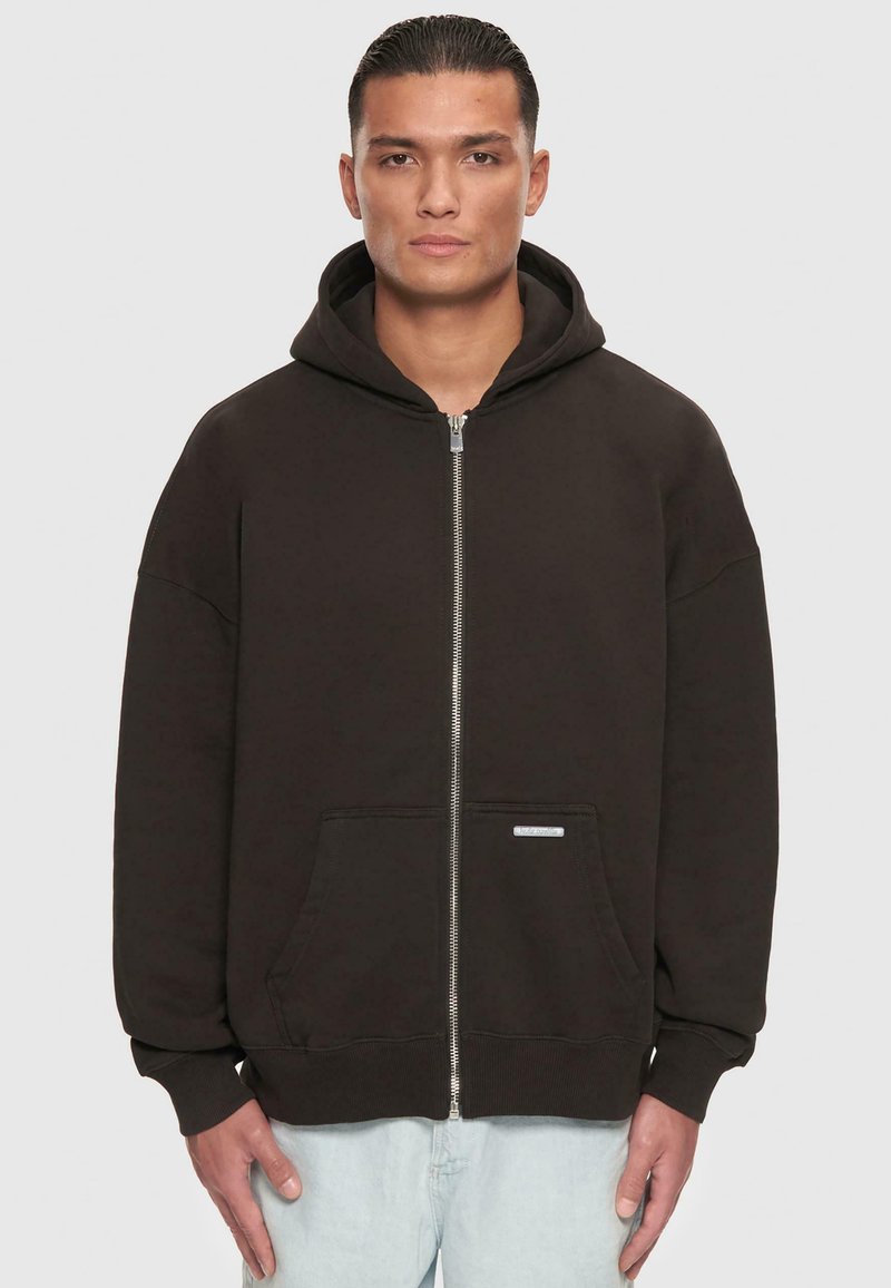 Schwarze Kapuzenjacke mit Reißverschluss aus Baumwollmischgewebe, ausgestattet mit einer Vorderstasche, einer Kordelzugkapuze und gerippten Bündchen. Minimales Branding-Details vorhanden.