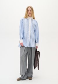 Šviesiai mėlynos juostelės marškiniai su baltais rankogaliais, pilkos plačios kojos kelnes ir ruda rankinė. Medžiaga atrodo lygi, audinys turi struktūrinį dizainą.