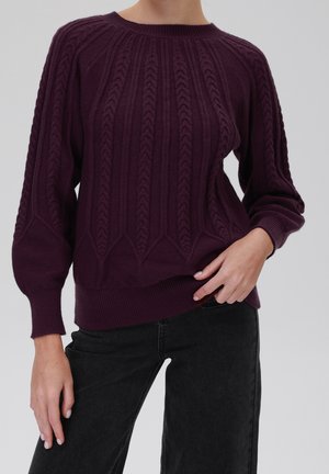 Femme portant un pull torsadé violet foncé à manches ballon et un jean noir, debout devant un fond clair et uni.
