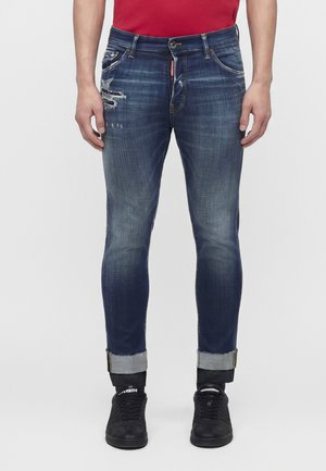 Smaltsiddende, udvaskede blå jeans med opkavlede bukseben, slidte detaljer på venstre lår, båret med sorte sneakers og en rød skjorte.