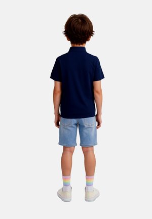 Bambino in piedi di spalle, indossa una polo blu navy, pantaloncini di jeans chiari, calzini a righe arcobaleno e sneakers bianche su uno sfondo semplice.