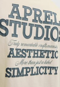 Tricou din bumbac crem, cu text proeminent de culoare gri închis: "APRELL STUDIOS," cu fraze decorative în fonturi și stiluri variate.