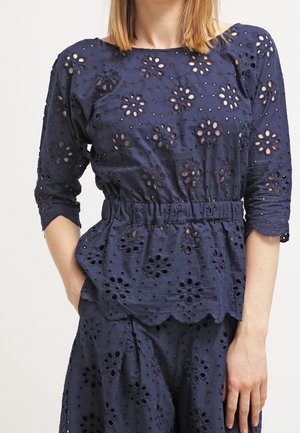 Vrouw die een marineblauwe blouse en broek van oogjesstof draagt met bloemmotieven in de uitgesneden patronen, driekwartmouwen en een elastische tailleband.